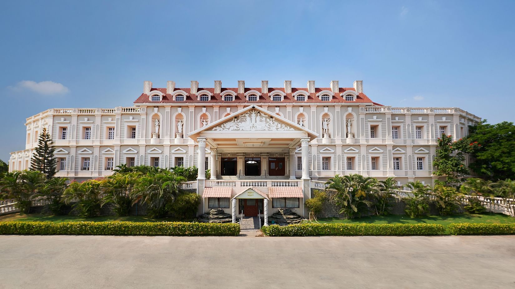Grand white facade with red gabled roof and columned portico, framed by lush greenery - Jenneys Residency, Coimbatore