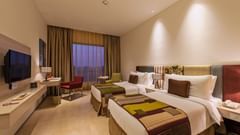 A deluxe twin room with plush beds, bed side table, flat screen TV, lamps, work desk, chair, and decorative items - Pride Elite Phaltan