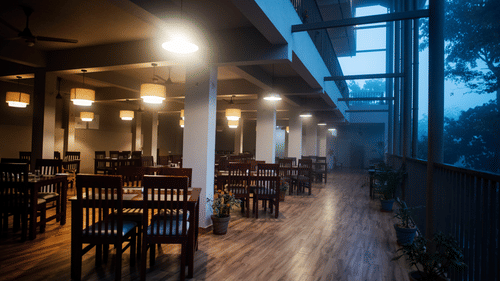A restaurant interior features wooden tables and chairs with pendant lights, overlooking a view.- Abad Brookside Lakkidi, Wayanad