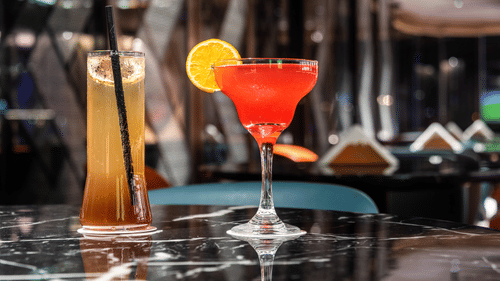Close-up of a cocktail glass with a vibrant red drink garnished with orange and cherry, set on a reflective surface at Asiatic Rooftop Bar and Restaurant.