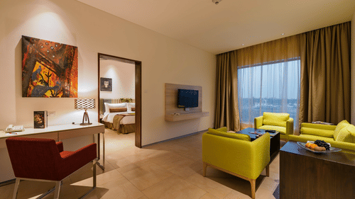 An image of the separate living room of the Suite with sofa set, centre table, work desk, chair, flat screen TV, and an expansive window with curtains - Pride Elite Phaltan