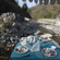 A table set with 2 plates and drinks under a canopy next to a rocky riverbed with hills.