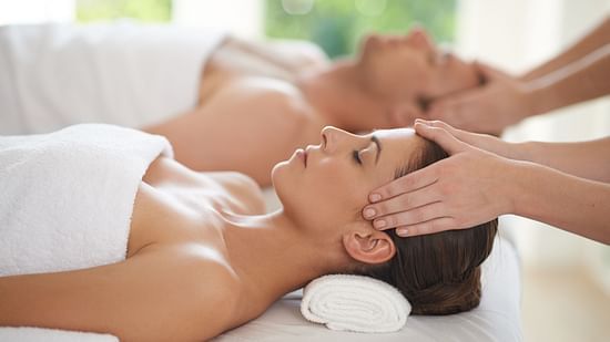 A couple laying on two white massage beds, wearing white towels and facing upwards, while their face is being massaged by two practitioners.