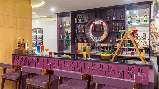 Bar counter lined with bottles, decorative wreath and purple upholstered stools at Mango Grove Hotel.