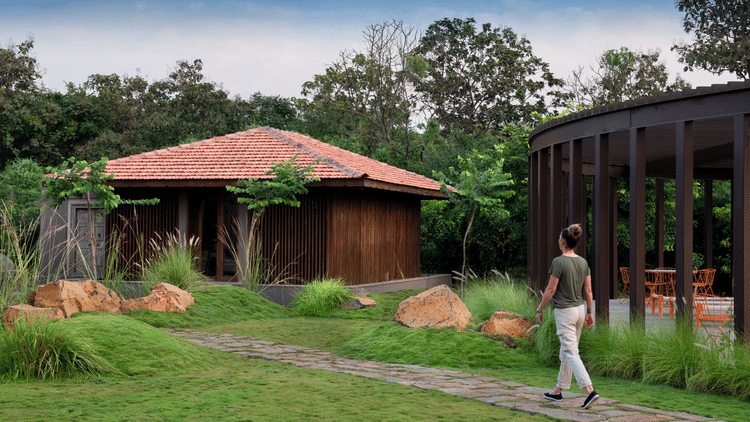 A person walking along a stone pathway surrounded by lush landscaped gardens at Aramness, with a wooden villa featuring a terracotta-tiled roof and a modern glass-walled pavilion nestled amid greenery and trees in the background.