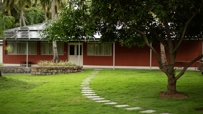 Lush green lawn with a curved stone pathway and cottages surrounded by trees at Ibex River Resort, Pollachi.