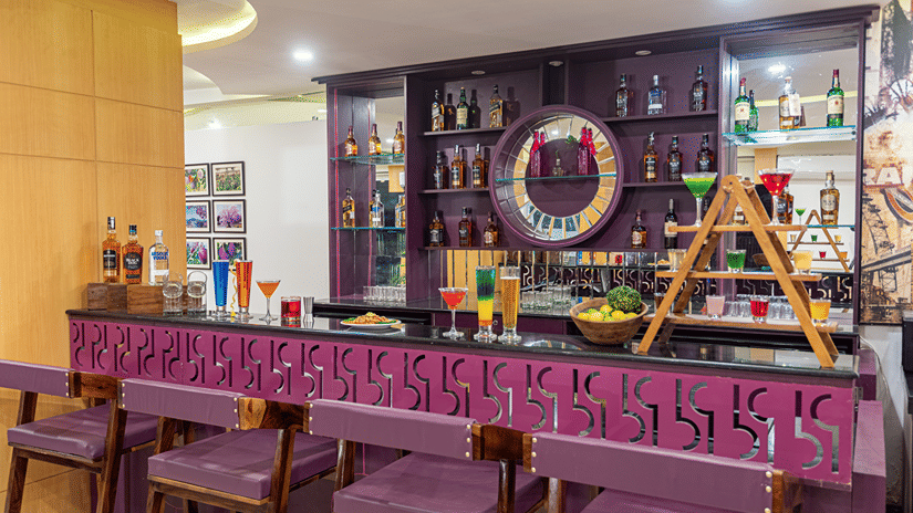 Bar counter lined with bottles, decorative wreath and purple upholstered stools at Mango Grove Hotel.
