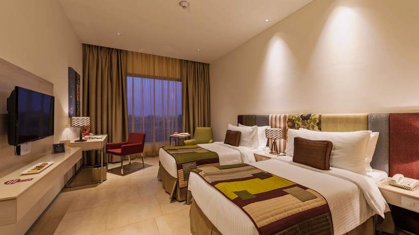 A deluxe twin room with plush beds, bed side table, flat screen TV, lamps, work desk, chair, and decorative items - Pride Elite Phaltan