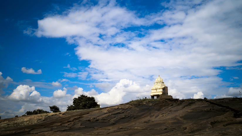A rocky terrain leading up to a small white temple structure under a wide blue sky with scattered clouds