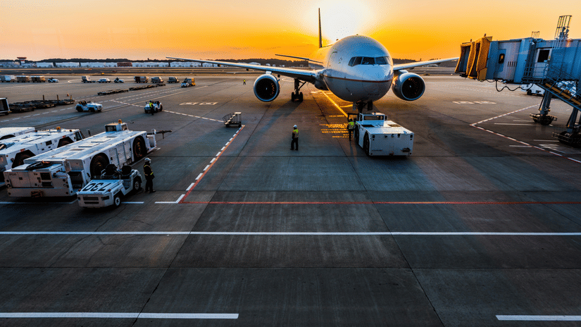 An aeroplane on the runway of an airport surrounded by other vehicles against a orange sky with the sun setting.