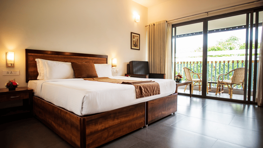 Bright bedroom at Abad Green Forest, one of the finest cottages in Thekkady, featuring wooden furnishings, crisp white bedding, and a balcony overlooking lush greenery.