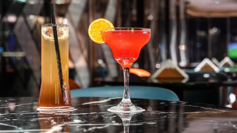 Close-up of a cocktail glass with a vibrant red drink garnished with orange and cherry, set on a reflective surface at Asiatic Lounge.