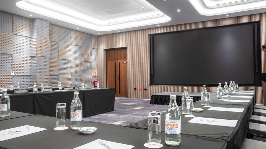 A modern conference or meeting room setup with long tables, bottled water, glasses, and notepads facing a large presentation screen.