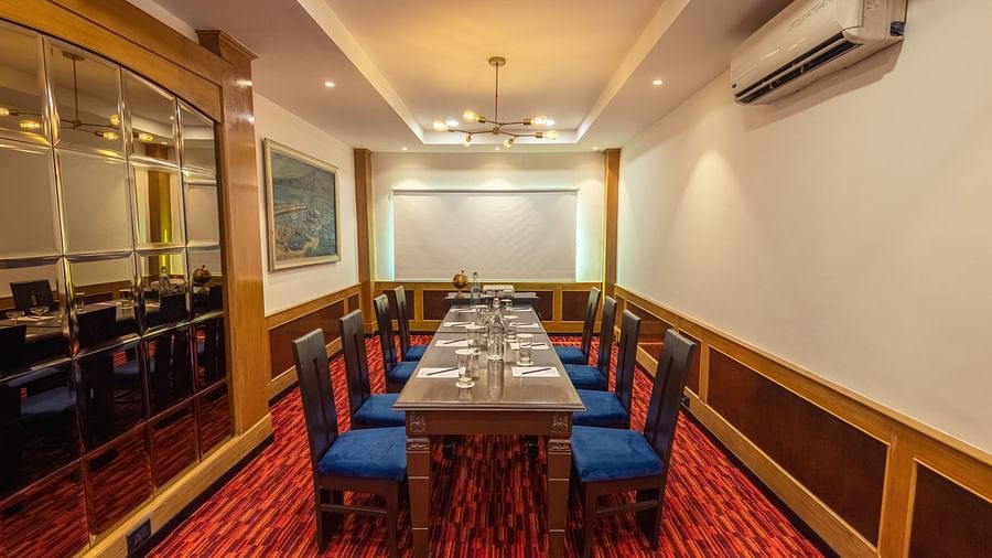 A private dining room with a long wooden table set for a meal, blue upholstered chairs, red patterned carpet, wood paneling, mirrored wall, and a modern chandelier.