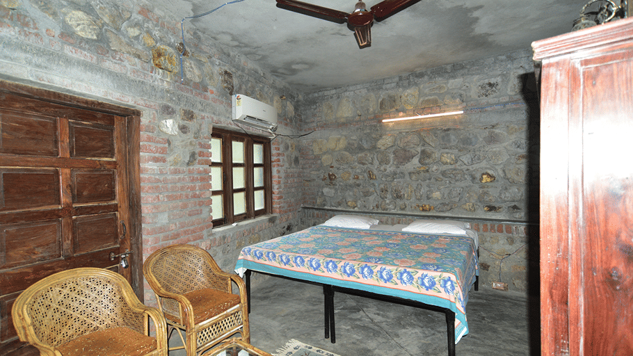 The Bird's Eye View Eco Cottage bedroom with rustic stone walls, wooden windows, and wicker chairs at Wild Brook Retreat, Rajaji National Park.