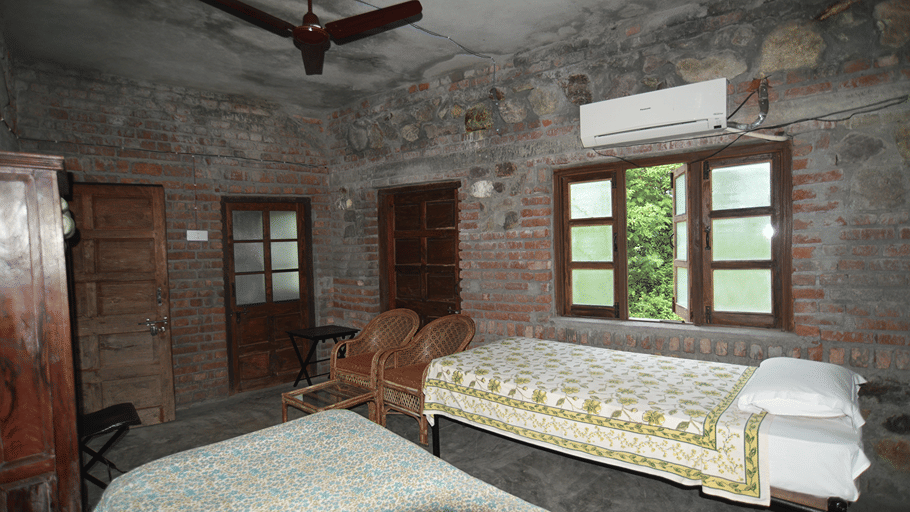 The Bird's Eye View Eco Cottage bedroom with rustic stone walls, wooden windows, and a double bed at Wild Brook Retreat, Rajaji National Park.