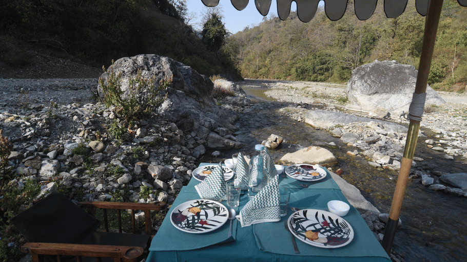 A table set with 2 plates and drinks under a canopy next to a rocky riverbed with hills.