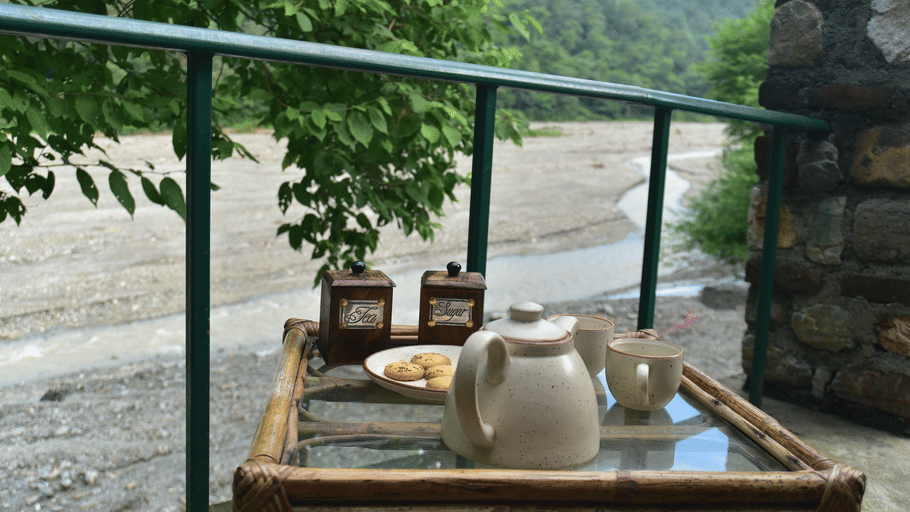 A teapot, 2 cups and snacks on a balcony table overlooking a wide river and green hills.