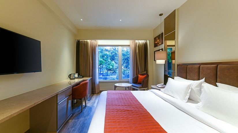A double bed room with a runner, pillows, seating area, work desk, TV and windows with curtains at Hotel Saket 27, New Delhi