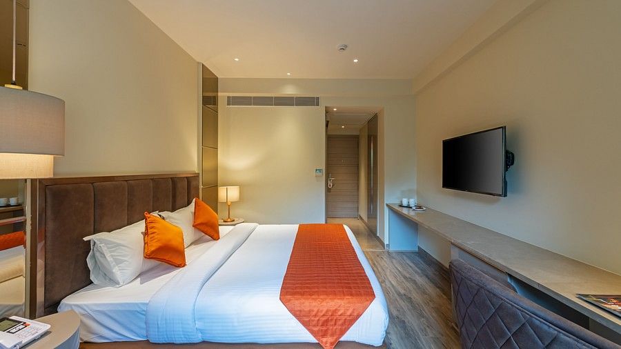 A side view of a double bed room with pillows, runner, TV, wooden floor, lamp and a hallway with a door at Hotel Saket 27, New Delhi
