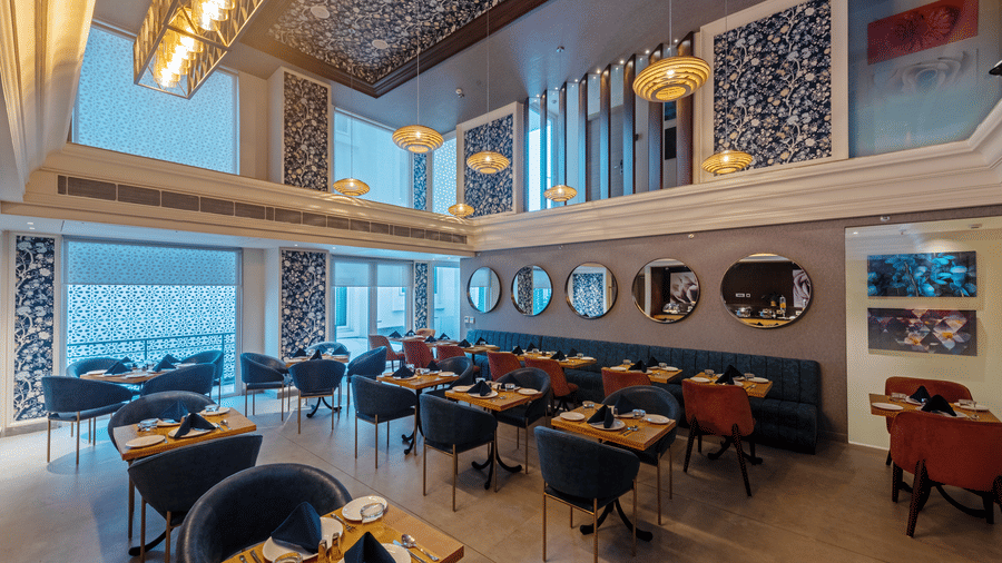 A side view of restaurant dining area with large windows, chairs, artistic wall décor, and ambient ceiling lights at Hotel Saket 27, New Delhi