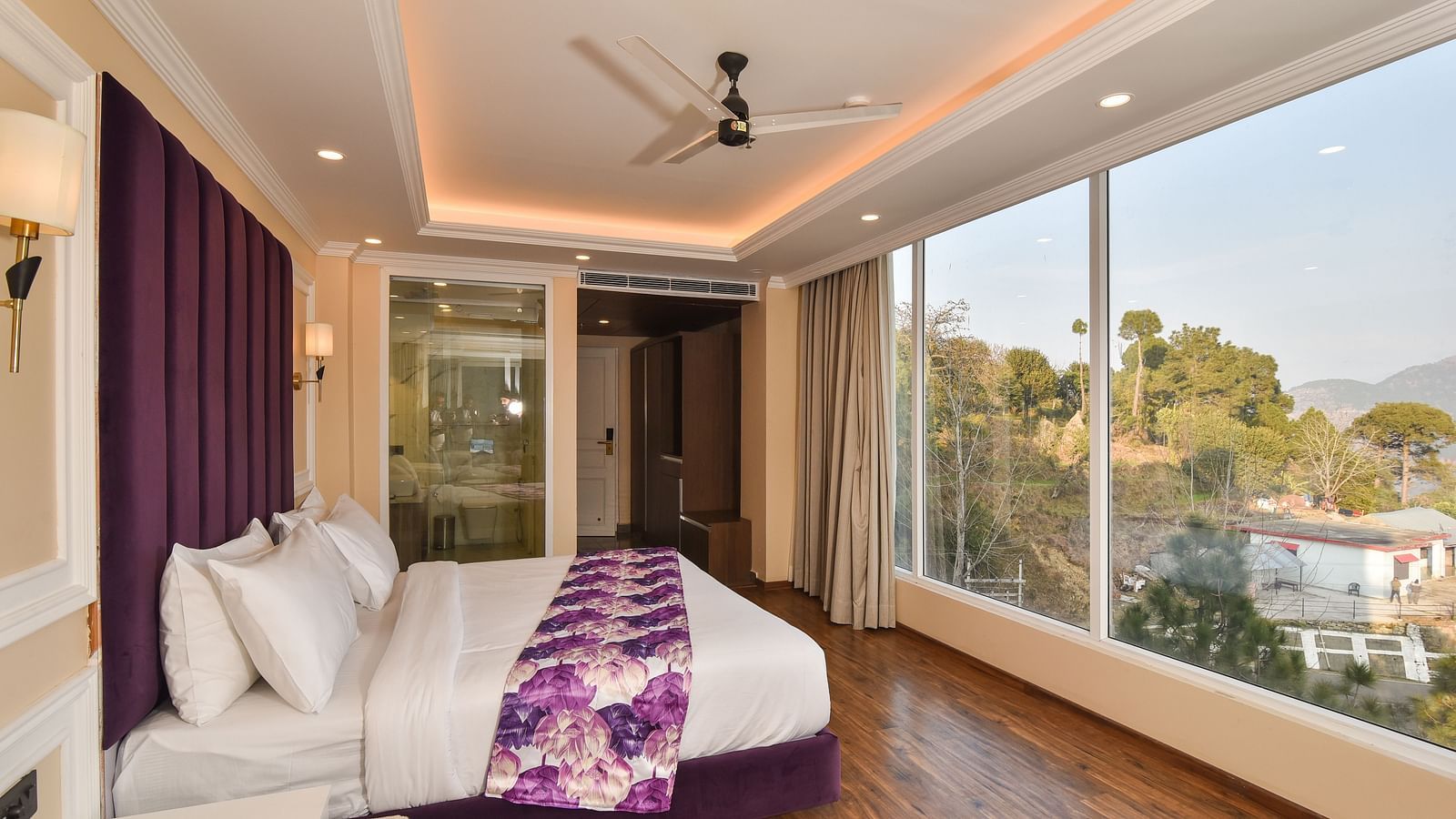 A well-designed bedroom with a large bed and purple bedding, large windows offering scenic views of the hills, and contemporary decor. The room has a relaxing and inviting atmosphere.