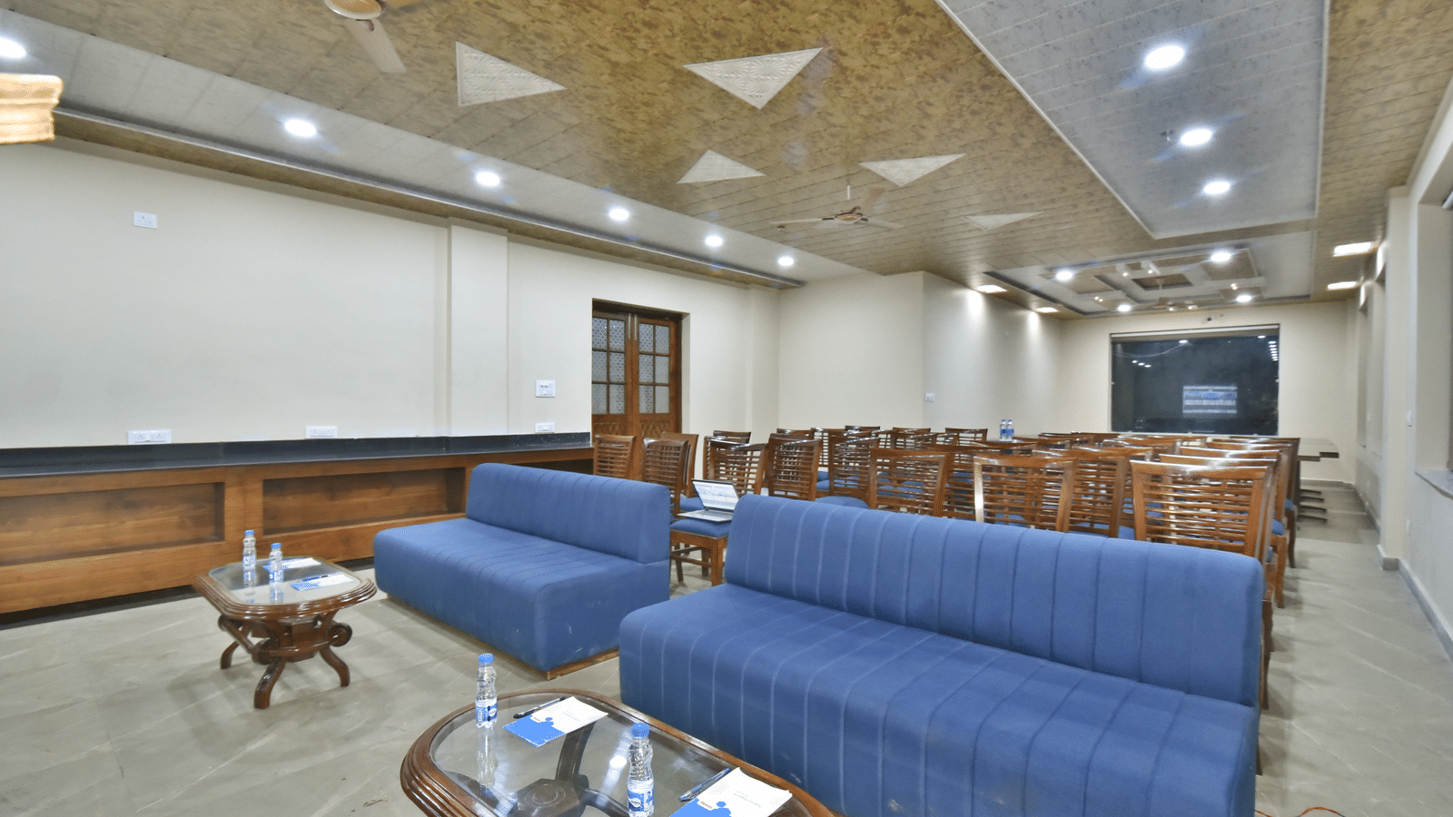 Meeting room at DLS Hotels with blue sofas, wooden chairs, and glass-top tables set with bottled water and notepads.