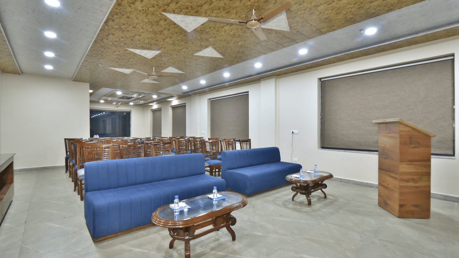 Conference hall at DLS Hotels with wooden chairs, blue sofas, podium, and ceiling fans under warm lighting.