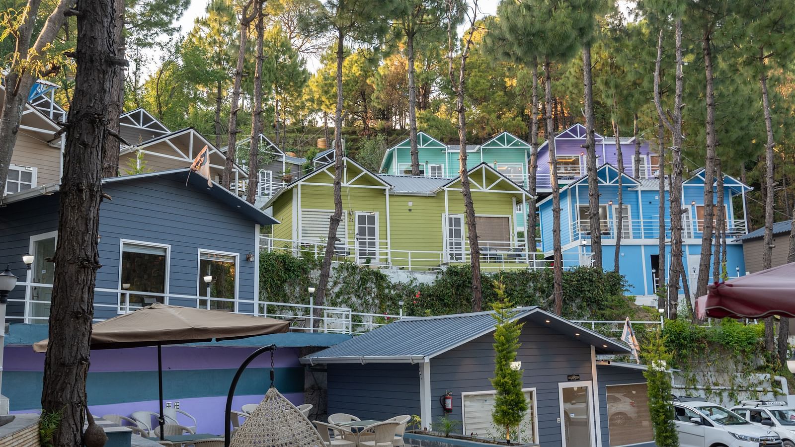View of colourful hillside cottages nestled among tall pine trees, with pastel blue, green, and purple facades, adjacent to a checkered courtyard and seating area below - DLS Nature Trinket Resort, Dalhousie.