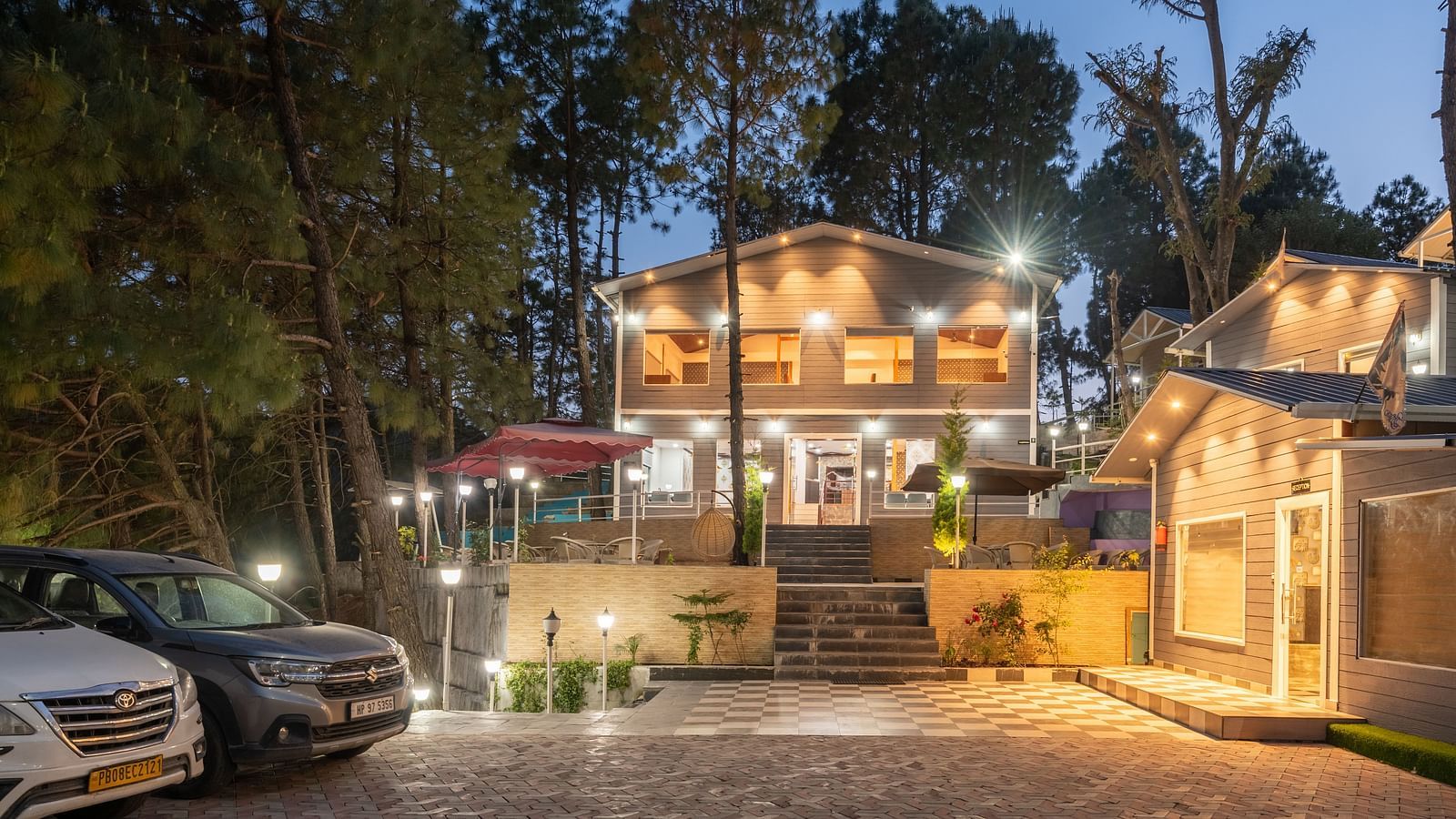 Evening view of a well-lit resort entrance with parked cars, a central staircase leading to a two-story building, and warm ambient lighting surrounded by tall pine trees - DLS Nature Trinket Resort, Dalhousie.