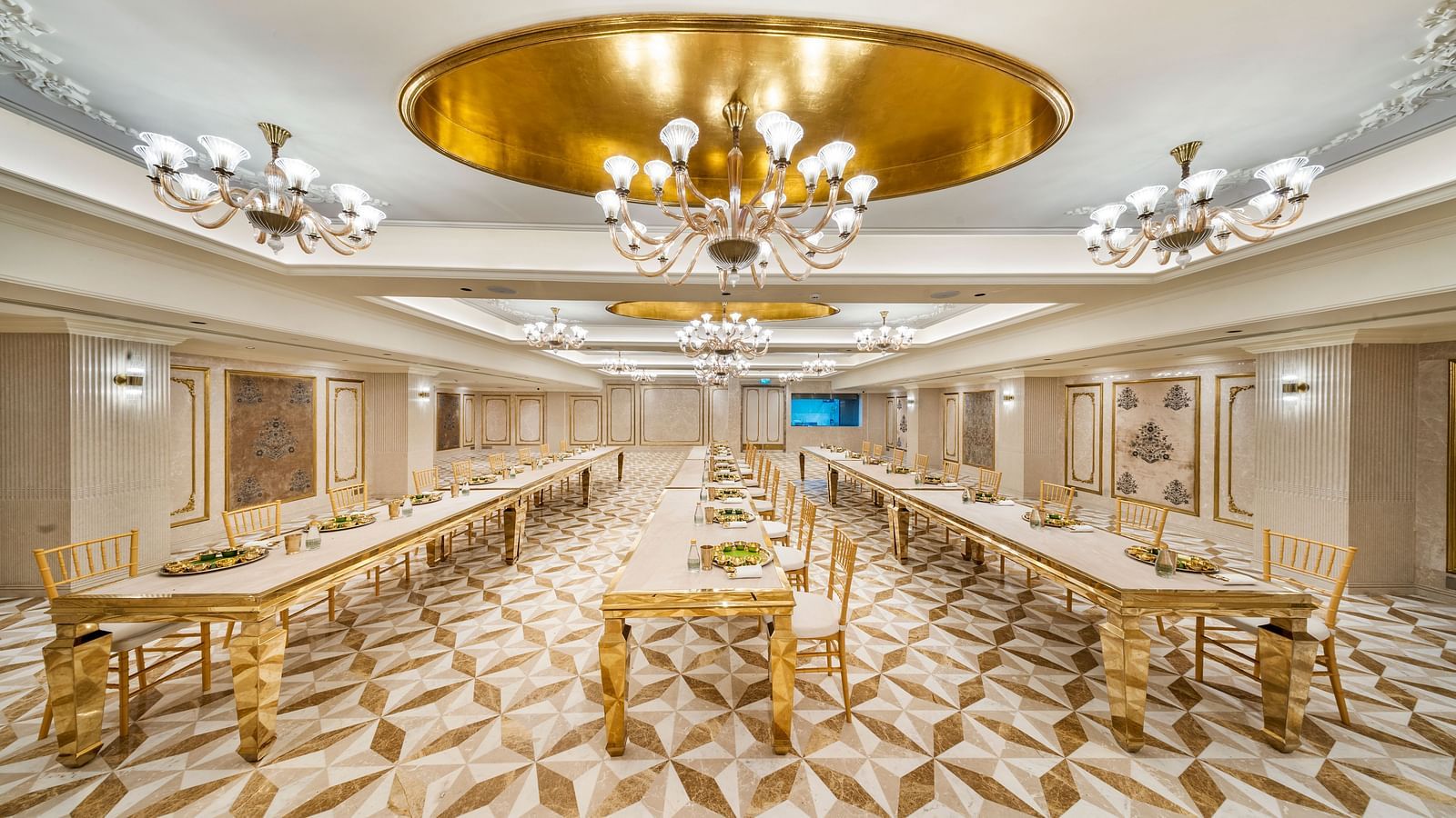 A lavish banquet hall at Grand Chennai by GRT Hotels, showcasing numerous golden tables and chairs under exquisite chandeliers.