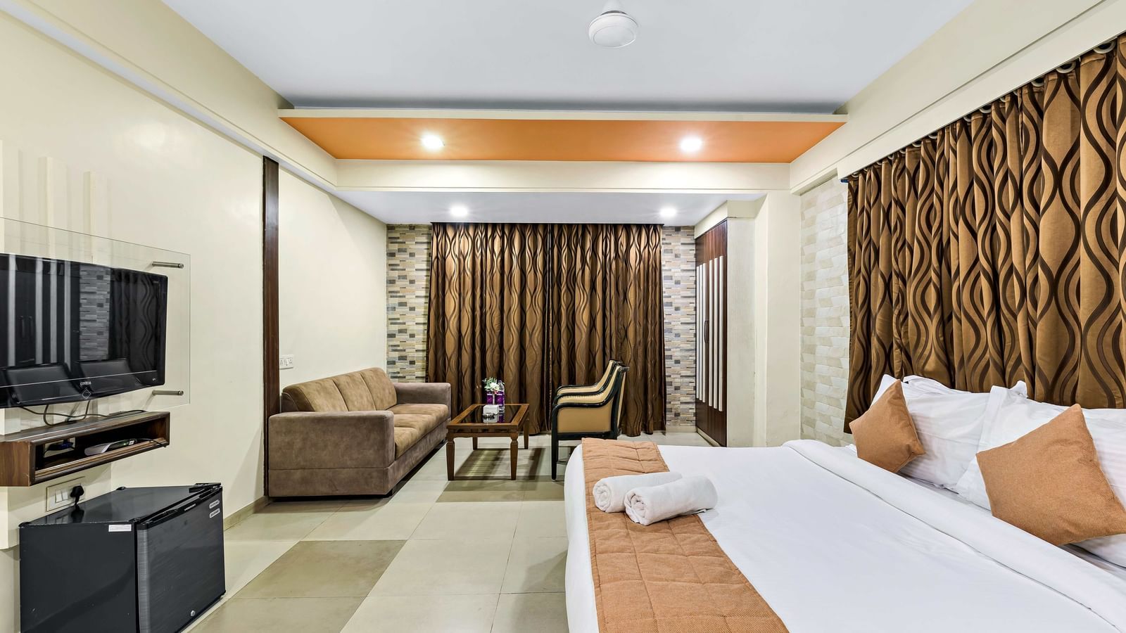 A well-lit suite in Krushnai Resort featuring a double bed with blue runner, a wall mounted TV, docking station, couch and a lounge chair.