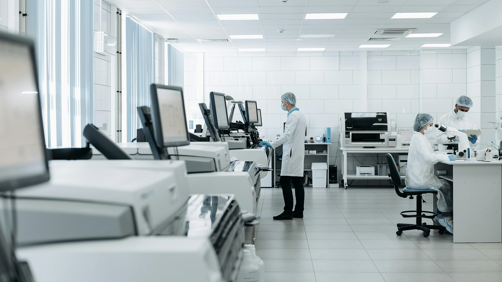 A well-lit laboratory setting shows scientists wearing personal protective equipment and conducting research with various equipment and computers. The lab features white walls and flooring, with several workstations equipped with monitors and scientific in