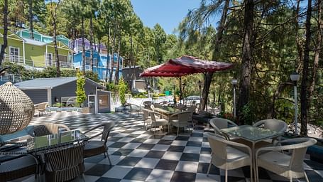 Outdoor dining area with checkered tile flooring, red umbrellas, and glass tables, surrounded by pine trees and colourful buildings - DLS Nature Trinket Resort, Dalhousie.