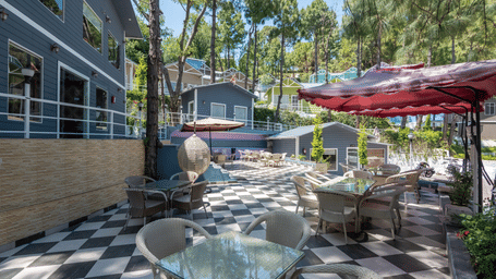 Sunny outdoor café area with glass tables, red umbrellas, and black-and-white checkered flooring, bordered by grey buildings and tall pine trees - DLS Nature Trinket Resort, Dalhousie.