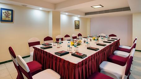 A formal boardroom set up with an elongated table covered in a red patterned cloth, surrounded by chairs at Lords Inn Vadodara.