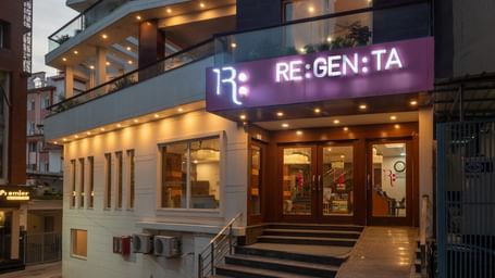 An eye-level view of the entrance to the Regenta Inn DLS, Rishikesh, in the evening. The hotel's brightly lit RE:GEN:TA sign is visible, along with the glass-paned doors of the lobby. The exterior walls are a mix of white and dark paneling, with warm light