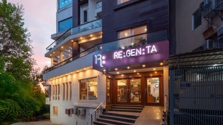 A vertical, low-angle shot of the multi-story Regenta Inn DLS, Rishikesh, taken at dusk. The building's modern architecture, with its dark paneling and large windows, is prominent. The bright RE:GEN:TA sign is positioned above the main entrance, and the su