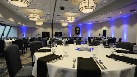 A wide view of a banquet hall set up with large, round tables, white linens, black napkins, and modern chandeliers. 