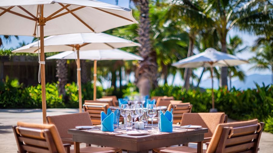 A picturesque dining setting under white umbrellas with set tables, palm trees and a lush background - Alibu Resorts Nha Trang