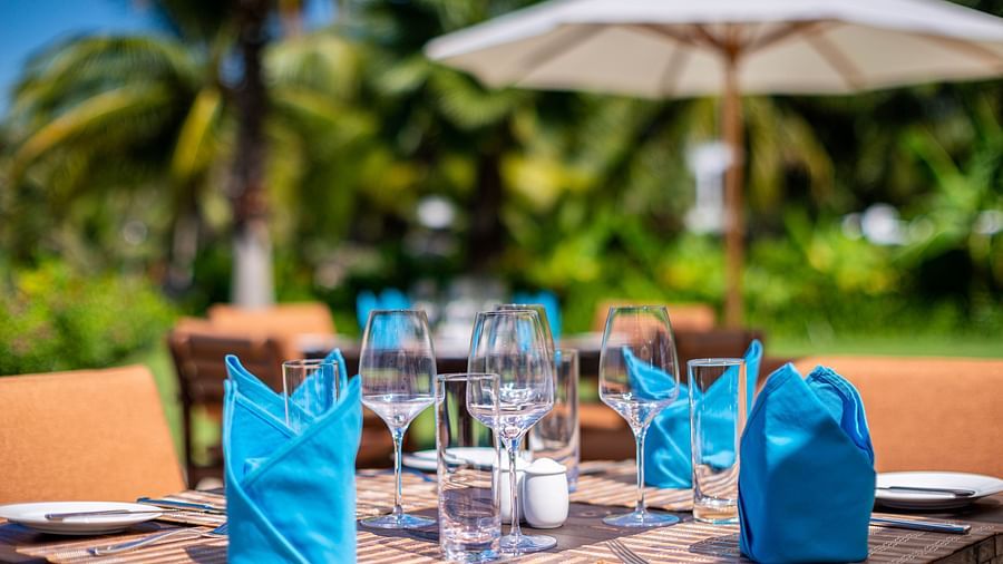 Close-up of an outdoor dining table with blue napkins, glasses and a white umbrella, with blurred palm trees in the background - Alibu Resorts Nha Trang