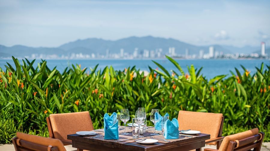 A seaside dining area with set tables under white umbrellas, vibrant plants and a panoramic view of the coastline and city - Alibu Resorts Nha Trang
