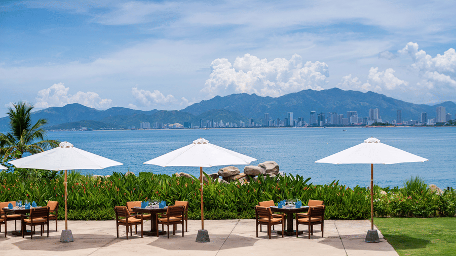 An alfresco dining setup with white umbrellas overlooking a serene bay and a distant cityscape at the best restaurant in Nha Trang inside Alibu Resorts.