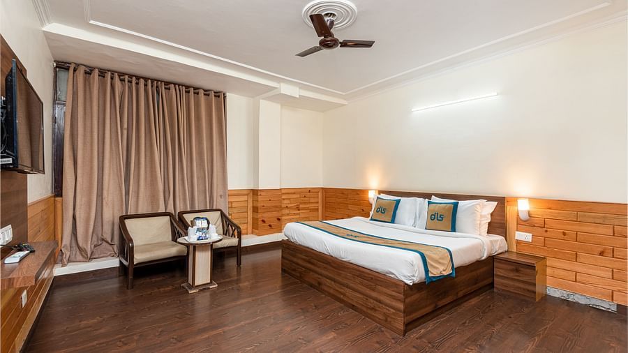 A well lit hotel room with a wooden floor, a large bed, two armchairs, a small table, and a ceiling fan | DLS Anupam Resort, Dharamshala