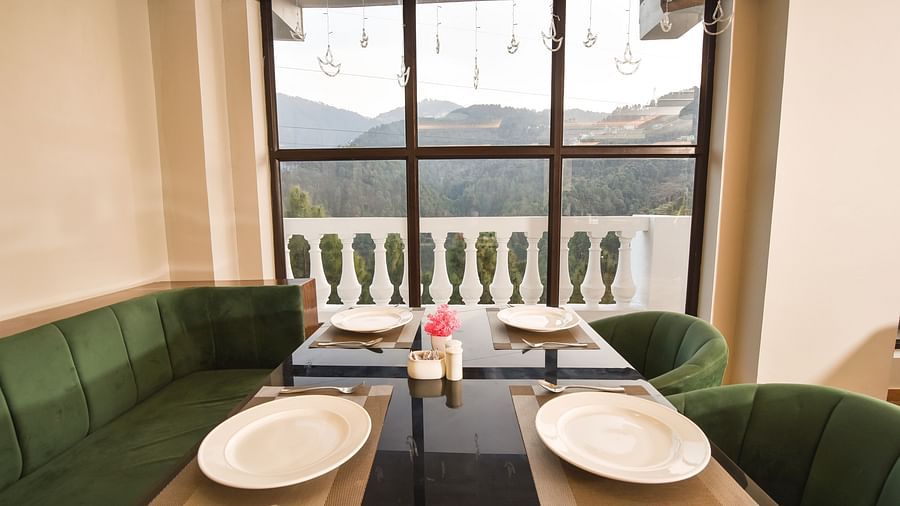 A cosy dining area with green upholstered chairs and a wooden table, offering panoramic views of the surrounding hills through large windows.