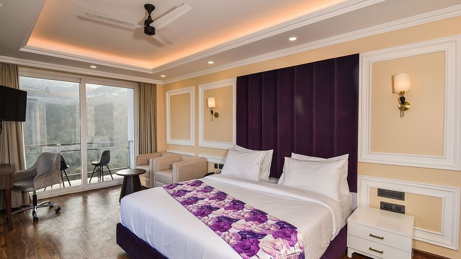 A vibrant bedroom with a large bed featuring purple accents and a bright atmosphere. The room includes large windows that provide scenic views of the hills outside.