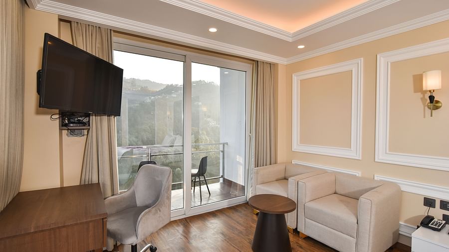 A modern bedroom with a king-sized bed, a seating area by the window, and large glass windows providing expansive views of the mountains.