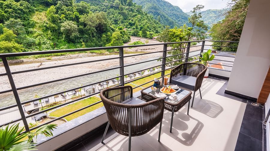 Overview of balcony of Premium Room at DLS Divine River Resort and  Spa featuring seating area with food kept on table and backdrop of river and forest views.