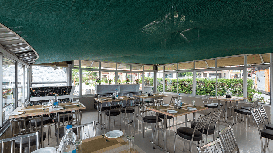 A cafe in Bhagsu with tables and chairs, featuring a green canopy overhead and large windows with views of the outside. The tables are set with plates, glasses, and bottles of water - DLS Meghavan Resort, Dharamshala