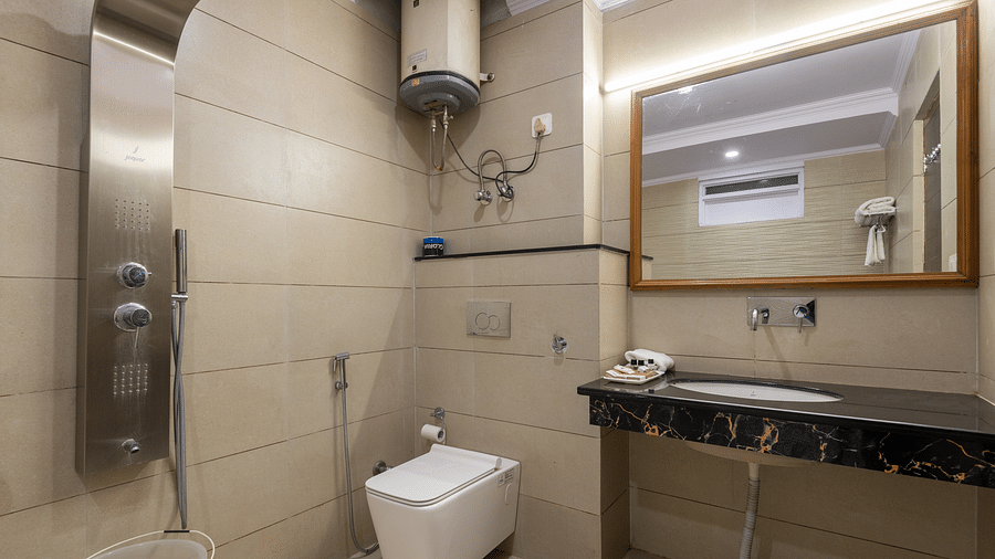 A bathroom with a shower panel, a modern toilet, a marble sink with toiletries, and a large mirror. The room also has a water heater mounted on the wall - DLS Meghavan Resort, Dharamshala